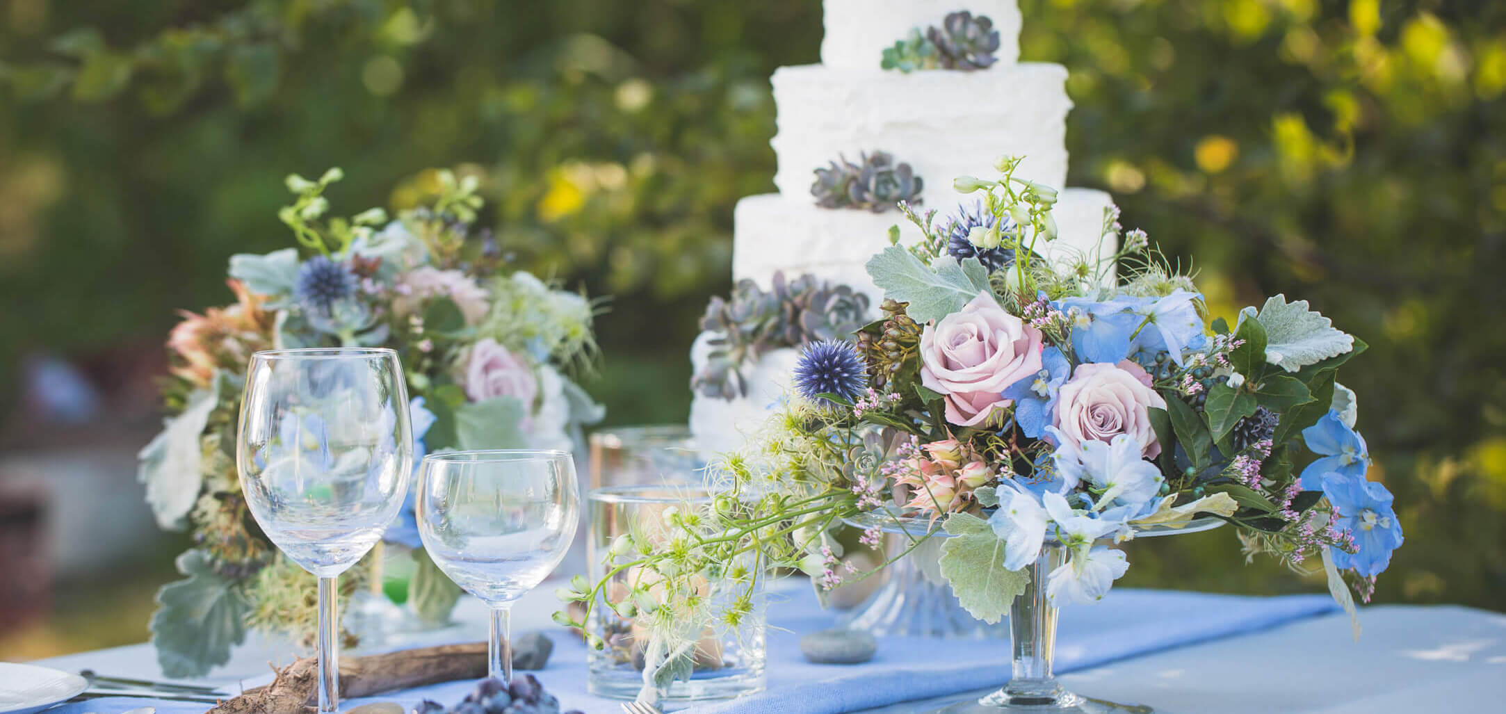 Sommerliche Tischdekoration (Kugeldistel, Silberblatt, Rittersporn, wilde Clematis, Antikrose) für eine Hochzeit mit Sokkulenten auf der Torte.